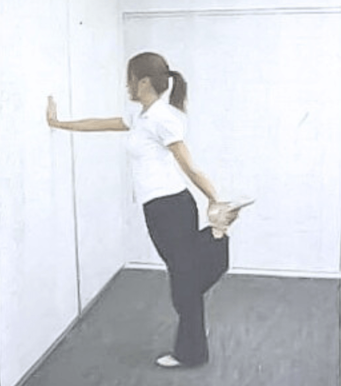 壁に片手をつき、反対の手で足首をつかんで太ももの前側を伸ばすストレッチを行っている様子