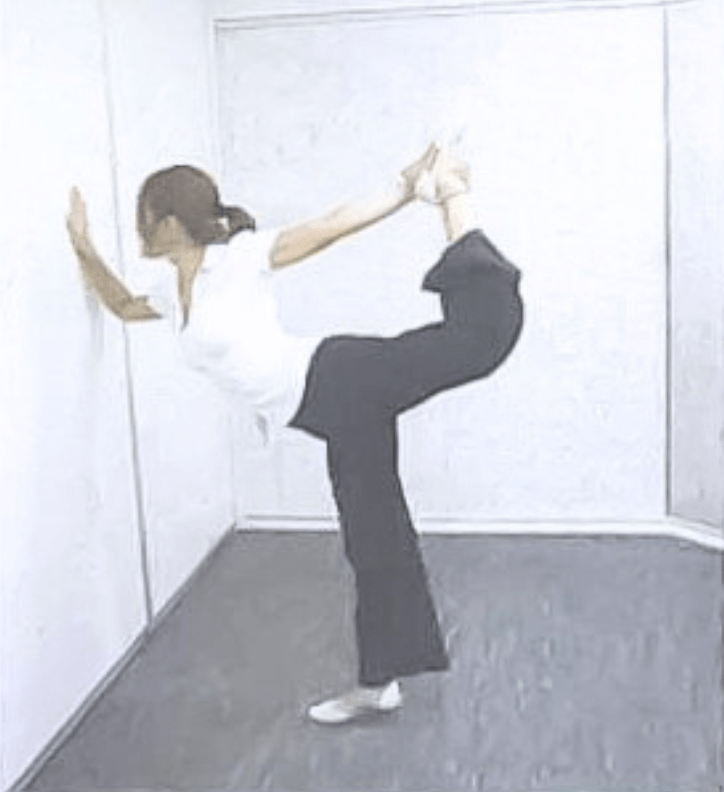 壁に片手をつき、反対の足を後ろへ大きく持ち上げてバランスを取りながら、ヨガのダンサーのポーズをとっている様子