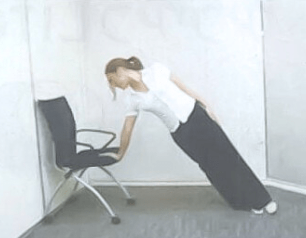 オフィスチェアに片手を添えて体をまっすぐに傾け、ヨガの賢者のポーズをアレンジしたストレッチを行う様子